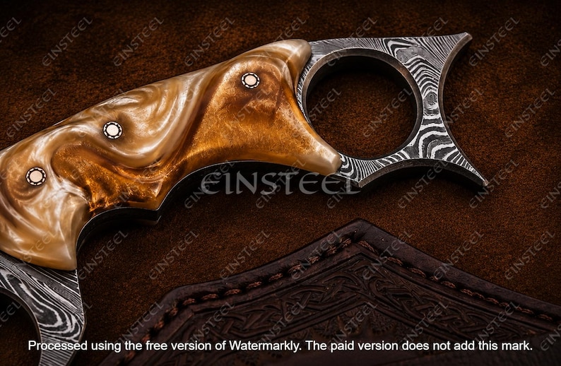 Pu&ograve; includere: Un coltello karambit con un manico in resina marmorizzata color ambra e crema con tre perni metallici circolari. La lama e l'anello presentano un distinto motivo ondulato in acciaio damasco. La parola EINSTEEL &egrave; visibile sul manico.