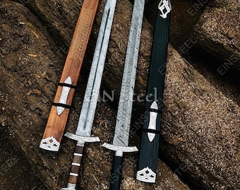 May include: Overhead shot of two swords with sheaths, resting on a wooden surface. The swords have silver blades with intricate patterns, ornate handles, and decorative crossguards. One sheath is brown, the other black, both with silver accents.
