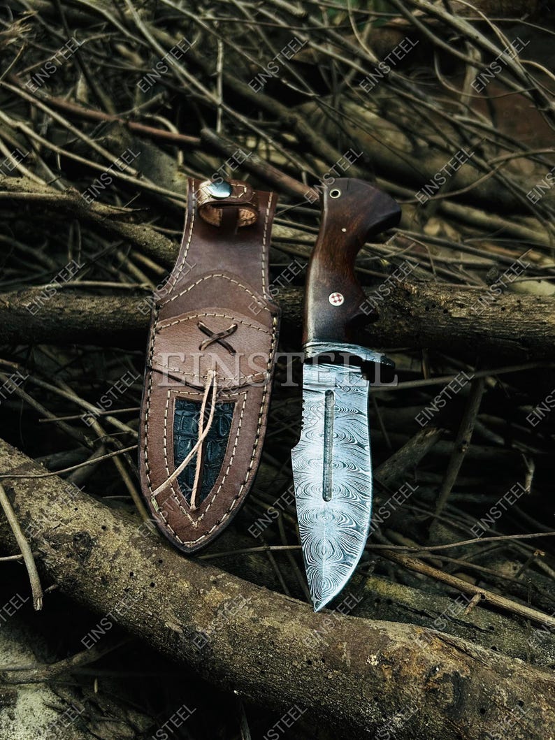 May include: A hunting knife with a patterned blade and a dark wooden handle rests beside its leather sheath. The sheath is brown with a decorative stitched design and a dark, textured panel. The knife blade has a unique, swirling pattern.