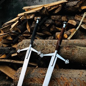 May include: Two long swords with silver blades and ornate crossguards. One sword has a black, twisted handle, and the other has a brown, wood-grain handle. The swords are displayed against a backdrop of stacked logs.