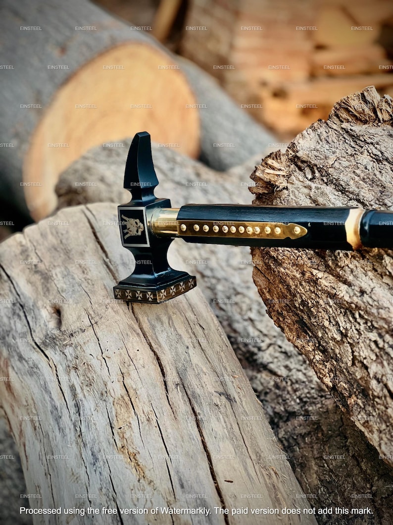 May include: A black and gold hammer with a decorative design. The hammer features a black handle with gold accents and a detailed head. The hammer is placed on a weathered wooden surface.