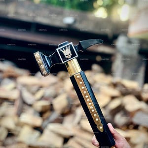May include: A black and gold hammer with a skull emblem on the head. The hammer has a black handle with gold accents and is held by a person. The hammer is designed for woodworking or construction.