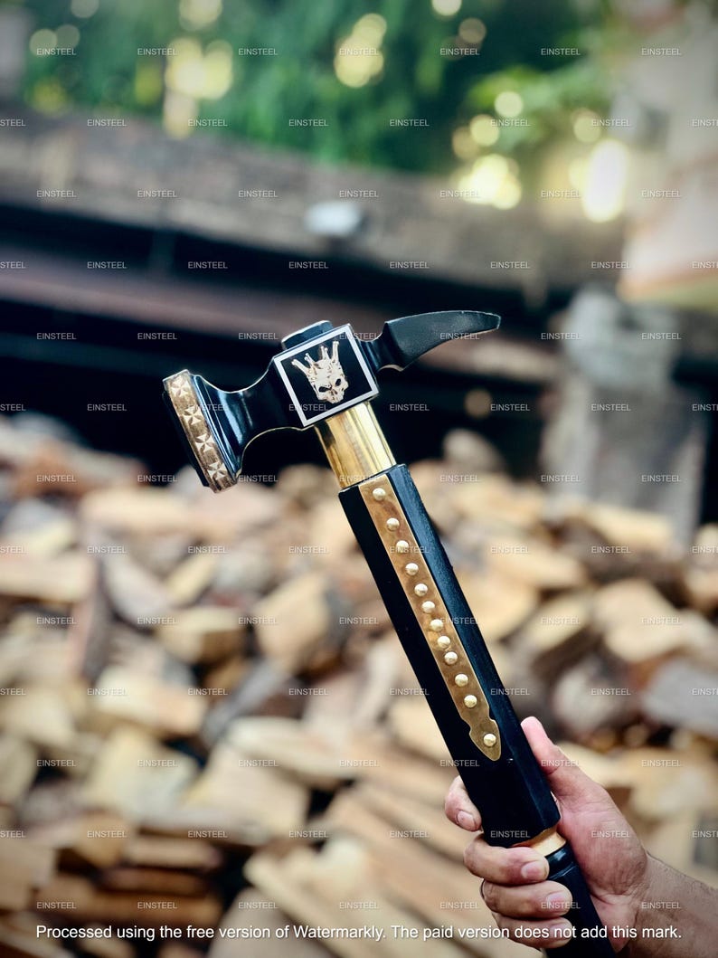 May include: A black and gold hammer with a skull emblem on the head. The hammer has a black handle with gold accents and is held by a person. The hammer is designed for woodworking or construction.