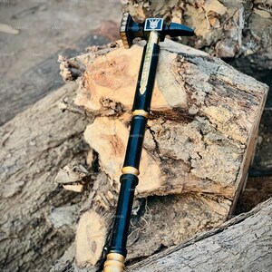May include: A black and gold hammer with a decorative handle. The hammerhead is square with a symbol in the center. The handle is black with gold accents and a gold base. The hammer is resting on a piece of wood.