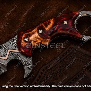 Pu&ograve; includere: Un coltello a lama fissa con una lama in acciaio damasco a motivi e un intarsio a zigzag in rame. Il manico &egrave; realizzato in resina lucida marmorizzata rossa e ambrata, fissato con tre perni metallici, e include un pomolo ad anello per il dito.