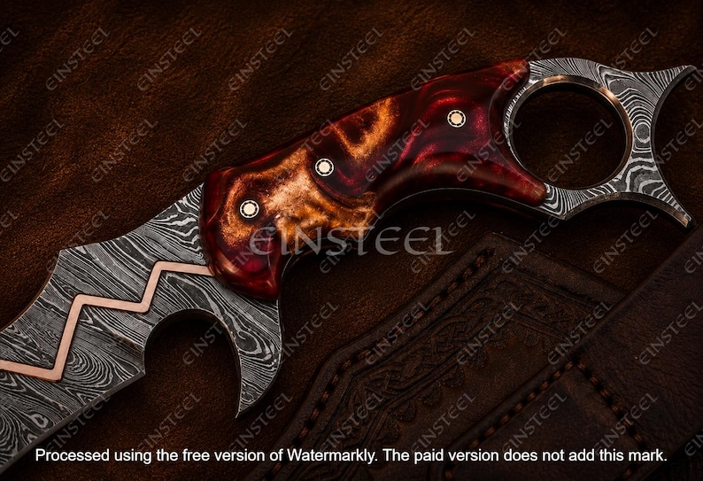 Pu&ograve; includere: Un coltello a lama fissa con una lama in acciaio damasco a motivi e un intarsio a zigzag in rame. Il manico &egrave; realizzato in resina lucida marmorizzata rossa e ambrata, fissato con tre perni metallici, e include un pomolo ad anello per il dito.