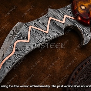 Pu&ograve; includere: Una lama di coltello curva con un distinto motivo in acciaio Damasco nero e argento con un intarsio centrale in rame a zigzag. Il manico &egrave; realizzato in materiale color ambra lucido con tre rivetti circolari, vicino a un fodero in pelle scura.