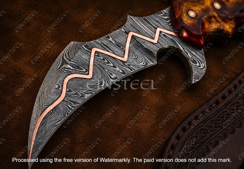 Pu&ograve; includere: Una lama di coltello curva con un distinto motivo in acciaio Damasco nero e argento con un intarsio centrale in rame a zigzag. Il manico &egrave; realizzato in materiale color ambra lucido con tre rivetti circolari, vicino a un fodero in pelle scura.