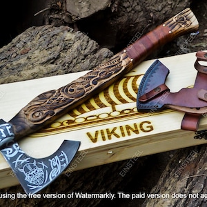 May include: A Viking axe with a wooden handle and a metal blade, resting on a wooden box. The axe head features a detailed design, and the box is engraved with the word "VIKING". A leather sheath is also included.