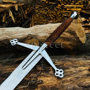 May include: A Scottish broadsword with a polished steel blade and a wooden handle. The hilt features a crossguard with decorative quatrefoil designs. The sword rests on a wooden log, with a background of stacked firewood.