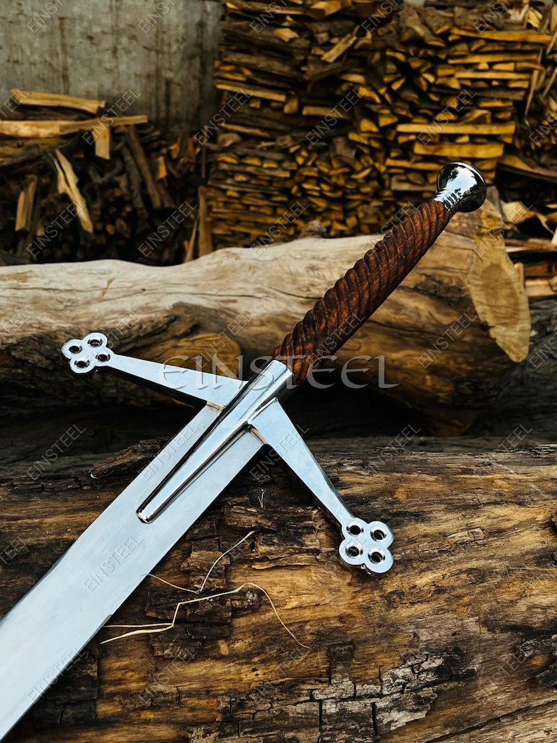 May include: A Scottish broadsword with a polished steel blade and a wooden handle. The hilt features a crossguard with decorative quatrefoil designs. The sword rests on a wooden log, with a background of stacked firewood.