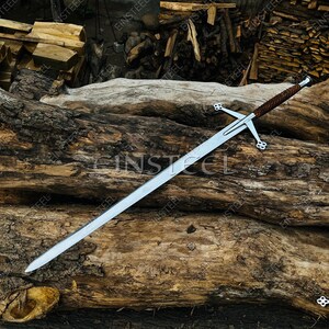 May include: A long, silver sword with a cross-shaped hilt and a brown leather-wrapped handle. The sword is resting on a weathered, textured log, with a background of stacked firewood. The hilt features decorative clover-shaped designs.