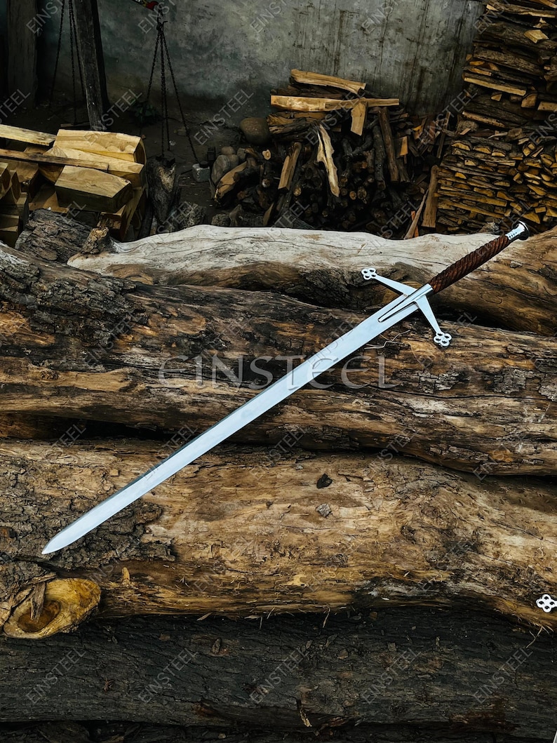 May include: A long, silver sword with a cross-shaped hilt and a brown leather-wrapped handle. The sword is resting on a weathered, textured log, with a background of stacked firewood. The hilt features decorative clover-shaped designs.