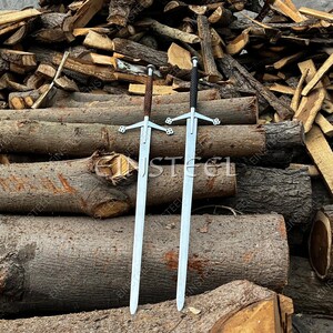 May include: Two medieval swords with silver blades and ornate crossguards. One sword has a brown leather-wrapped handle, the other a black handle. The swords are displayed against a backdrop of stacked logs and firewood.