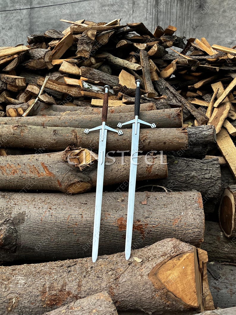 May include: Two medieval swords with silver blades and ornate crossguards. One sword has a brown leather-wrapped handle, the other a black handle. The swords are displayed against a backdrop of stacked logs and firewood.