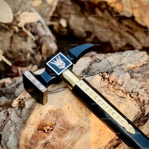 May include: A black and gold hammer with a decorative handle and a square head. The hammer features a brass handle and a silver emblem with a skull design. The hammer is resting on a wooden surface.