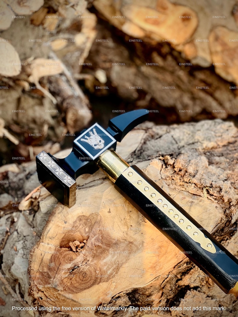 May include: A black and gold hammer with a decorative handle and a square head. The hammer features a brass handle and a silver emblem with a skull design. The hammer is resting on a wooden surface.