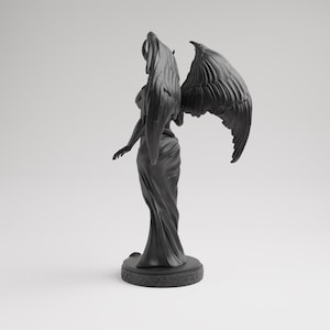 Op de afbeelding: Een zwart beeld van een gevleugelde figuur met hoorns, staand op een ronde basis. De figuur draagt een vloeiende jurk en heeft uitgestrekte armen. De vleugels zijn gedetailleerd en gespreid achter de figuur.