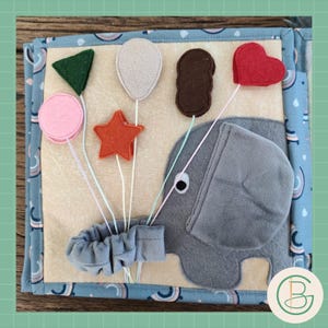 Op de afbeelding: Een zacht, kleurrijk stoffen activiteitenboek voor baby's en peuters. Het boek bevat een grijze olifant, vilten vormen en een blauwe rand met een regenboogpatroon. De vormen zijn onder meer een roze cirkel, een oranje ster, een groene driehoek, een witte ballon, een bruine rechthoek en een rode hart.