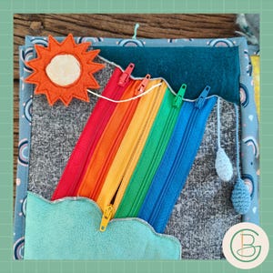 May include: A colorful felt activity book featuring a rainbow with zippers in red, orange, yellow, green, and blue. The book includes a felt sun, a cloud, and two crocheted rain drops. The background is a gray fabric with a blue border.