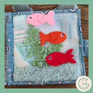 Puede incluir: Una página de libro de actividades de tela con una escena submarina. Peces de fieltro rosa, naranja y rojo nadan sobre algas verdes de ganchillo y tela azul esponjosa. El fondo es una tela estampada azul y blanca.