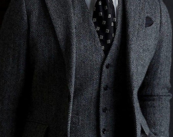 May include: A grey herringbone three-piece suit, comprising a blazer, waistcoat, and trousers, paired with a white collared shirt and a black tie with a white geometric design. A pocket square is visible in the blazer's pocket.