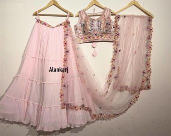 Könnte beinhalten: Ein mehrlagiger rosa Rock mit Blumenstickerei, ein passendes rosa Netz-Dupatta mit Blumenstickerei und eine rosa ärmellose bestickte Bluse. Die Bluse hat einen V-Ausschnitt und einen kleinen Quastenanhänger am Ausschnitt. Der Text "Alankari" ist auf dem Rock sichtbar.