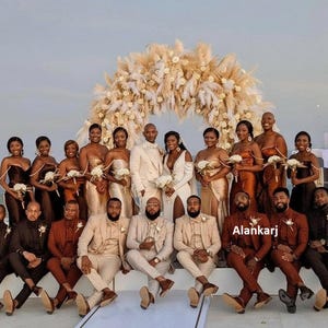 May include: A group of people, likely a wedding party, posing outdoors. The women wear various shades of gold and brown gowns, while the men are in suits of similar colors. A large floral arch is in the background.