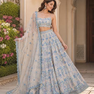 May include: A beige lehenga choli set featuring intricate blue floral embroidery and sequin detailing. The ensemble includes a sleeveless crop top with thin straps, a voluminous floor-length skirt, and a matching sheer dupatta with a scalloped edge.