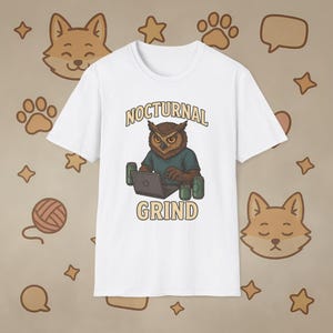 May include: White t-shirt with a cartoon owl wearing a teal shirt and working on a laptop. The text "NOCTURNAL GRIND" is above and below the owl illustration. Two green cans are on either side of the laptop.