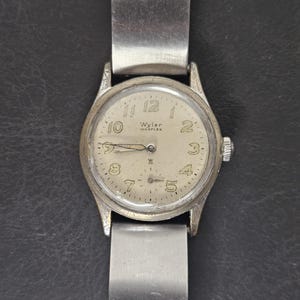 Puede incluir: Un reloj de pulsera vintage Wyler Incaflex con caja y correa plateadas. La esfera del reloj es de color crema con números y agujas negros. La correa es de metal cepillado.