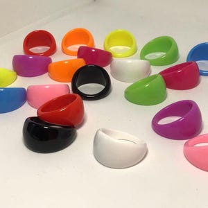 May include: Assortment of colorful plastic rings in various colors, including red, blue, green, yellow, black, white, and pink. The rings are all the same size and shape, and they are arranged in a semi-circle on a white surface.