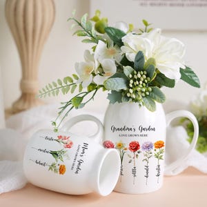 May include: Two white ceramic mugs with floral designs and the text "Mom's Garden Love Grows Here" and "Grandma's Garden Love Grows Here". The mugs are sitting on a white surface with a bouquet of white flowers in a white vase.