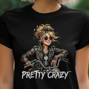 May include: Black t-shirt featuring a graphic of a woman on a motorcycle, wearing a leather jacket and sunglasses. The image includes the text "PRETTY CRAZY" in white. The woman has tattoos and a colorful headscarf.