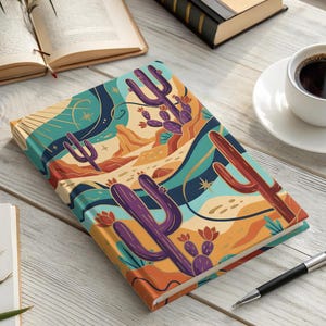 May include: A rectangular notebook featuring a colorful desert landscape illustration on the cover, depicting purple and red cacti, rolling sand dunes, and a stylized sun against a teal and orange background, resting on a light wood surface.