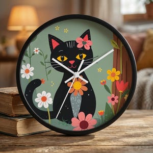 Könnte beinhalten: Eine runde, schwarze Uhr mit einer Illustration einer schwarzen Katze mit Blume, umgeben von bunten Blumen auf grünem Hintergrund. Die Uhrzeiger sind weiß. Die Uhr steht auf einer Holzoberfläche.