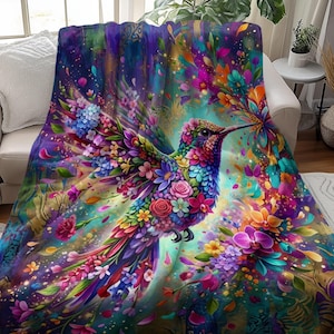 May include: A colourful blanket with a hummingbird design. The bird is decorated with flowers in shades of pink, purple, blue, and yellow. The background has a watercolour effect with similar colours. The blanket is draped over a white sofa.