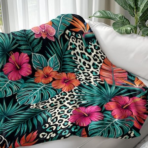May include: A throw blanket with a tropical floral and animal print design. The blanket features large pink and orange hibiscus flowers, green leaves, and a leopard print pattern on a black background. The blanket is draped over a white sofa.