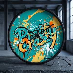 May include: A round wall clock with a black frame. The clock face features the words "Pretty Crazy" in a teal and orange graffiti style against a teal background with white and orange paint splatters. Black eyeglasses rest in front of the clock.