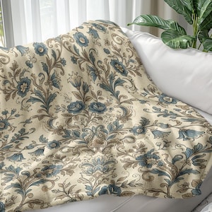 May include: A beige throw blanket with a repeating floral pattern in shades of blue and brown, draped over a white sofa. The blanket features a detailed, vintage-inspired design with stylized flowers and leaves.