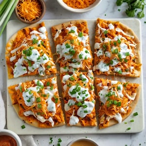 May include: Overhead shot of six flatbread pizza slices. Each slice is topped with orange shredded chicken, white sauce, and green onions. The flatbreads are arranged on a white rectangular serving board, with bowls of ingredients in the background.