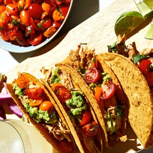 May include: Four soft tacos filled with shredded chicken, guacamole, diced tomatoes, and cilantro. The tacos are on a wooden cutting board with a bowl of salsa and limes in the background.