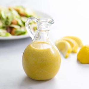Puede incluir: Una botella de vidrio transparente con un líquido amarillo en su interior. La botella tiene un asa y un cuerpo redondo. La botella está sobre una superficie blanca con rodajas de limón cerca.