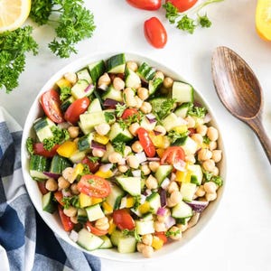 Puede incluir: Un bol blanco lleno de una ensalada colorida de garbanzos, pepinos, tomates, cebolla roja y pimientos amarillos. La ensalada está adornada con perejil fresco.