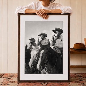 May include: A black and white framed wall art print depicting three cowboys wearing wide-brimmed hats, each sitting on a horse and drinking from a white mug. The print is encased in a dark brown wooden frame with a white mat border, showcasing a scene.