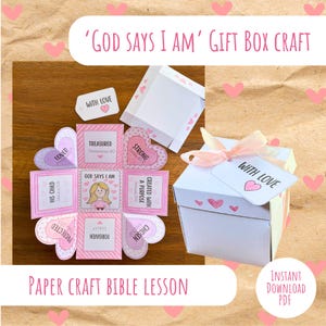 Puede incluir: Una caja de regalo blanca con una cinta rosa y una etiqueta que dice "Con amor". La imagen presenta manualidades de papel rosa y blanco con frases como "Dios dice que soy" y "Atesorado". También se ve el texto "Lección bíblica de manualidades".