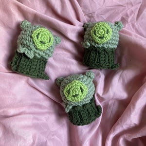 May include: Three handmade crochet piglet dolls. Each piglet has a light green face with a smiling face, gray ears, and a dark green body. The dolls are displayed on a pink fabric background.