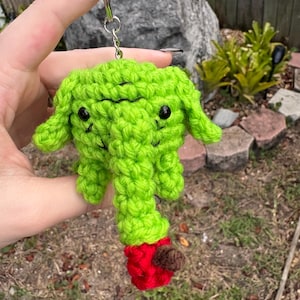 May include: A bright lime green crocheted elephant keychain featuring black bead eyes and a small red apple with a brown stem held at the end of its trunk. The stuffed animal is attached to a silver metal split ring and chain for hanging.