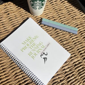 May include: A white spiral bound journal with the text "Make Progress: The Running Journal" in green and black. The text "Run, Review, Reflect" is also in green and black. A green and black running figure is in the bottom right corner.