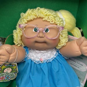 May include: A Cabbage Patch Kids doll with blonde curly hair and pink-rimmed glasses. The doll is dressed in a blue dress with a white lace collar and a small pink rose. A tag with the Cabbage Patch Kids logo is visible.